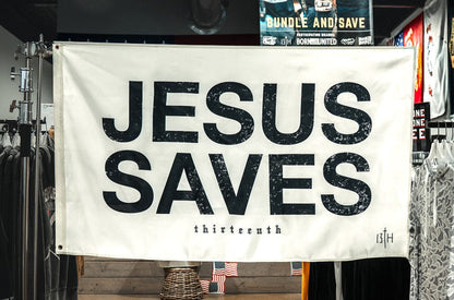 The 13th Jesus Saves Flag - Ivory hangs boldly in a clothing store, its striking black text standing out among racks of clothes and promotional signs for a powerful statement of faith.