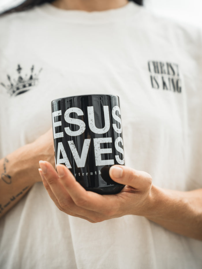 A tattooed person holds the 13th Jesus Saves Mug, wearing a white "CHRIST IS KING" shirt with a crown graphic. The bright, blurred background highlights the product.