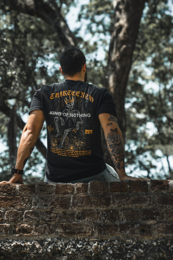 Person wearing a black t-shirt with graphics and text, standing on a stone wall with trees in the background.