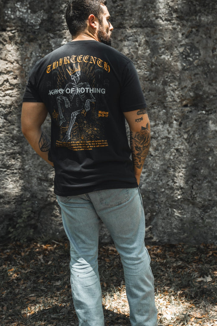 A man with short dark hair and tattoos stands outdoors in 13th's King of Nothing Premium Tee—a black graphic shirt—paired with light blue jeans, sunlight filtering through trees onto a stone wall.