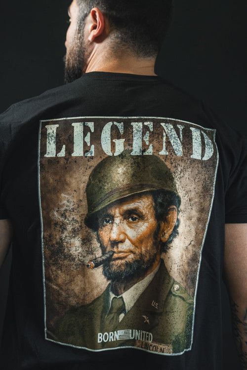 A tattooed man faces away, wearing Born United Private Label’s "Legend - Lincoln" tee, featuring Abraham Lincoln in military gear smoking a cigar with "LEGEND" above him—a tribute to American heritage.