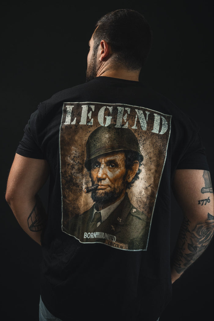 A man faces away, wearing the Legend - Lincoln tee by Born United Private Label, featuring Abraham Lincoln as a soldier with a cigar under "LEGEND" in bold. The dark shirt highlights his tattoos and celebrates American heritage.