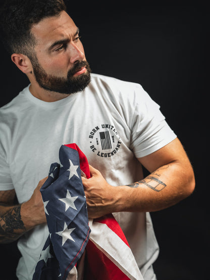 A man with a beard and tattoos holds a folded American flag. He is wearing a white T-shirt that reads BORN UNITED, BE LEGENDARY. The background is dark, highlighting the subject and the flag.