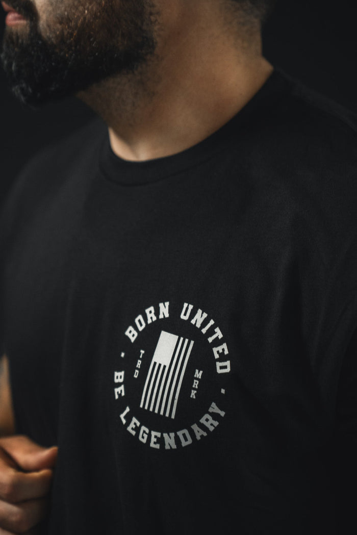 A person wearing the Legend - Washington tee by Born United Private Label, featuring a circular white "BORN UNITED BE LEGENDARY" logo and stylized flag—ideal for those proud of American heritage. Only their lower face and upper torso are shown.