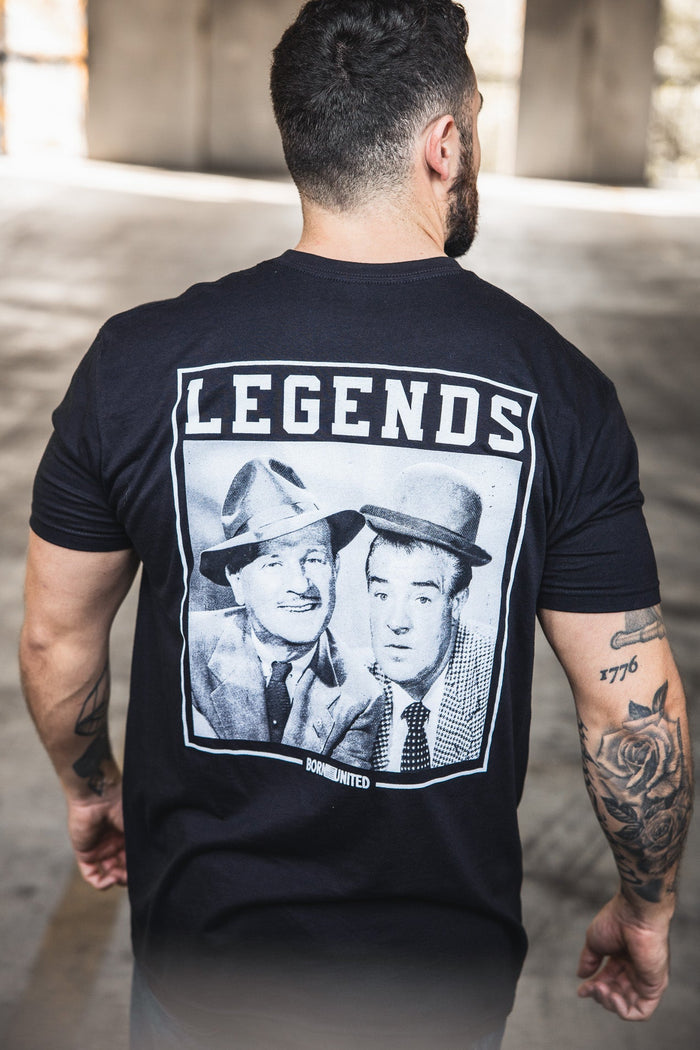 A tattooed man stands with his back to the camera, wearing a Born United Private Label "Legends - Bud and Lou" black T-shirt featuring vintage photos of two men in suits and hats, with LEGENDS printed above them.