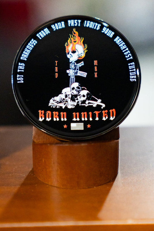 The Let the Darkness Challenge Coin by Born United Private Label features a round black tin with a flaming skull, crossed rifles, and central skulls. The tin reads “LET THE DARKNESS FROM YOUR PAST IGNITE YOUR BRIGHTEST FUTURE” and “BORN UNITED,” plus an American flag.