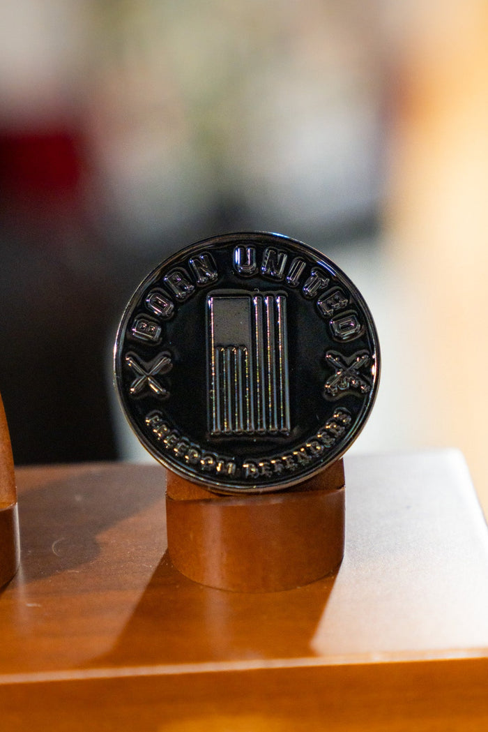 The Let the Darkness Challenge Coin by Born United Private Label is a black circular coin featuring a vertical column and crossed tools. Marked "IRON UNITED" and "STRENGTH BROTHERHOOD," it stands upright as a powerful symbol on a wooden surface.