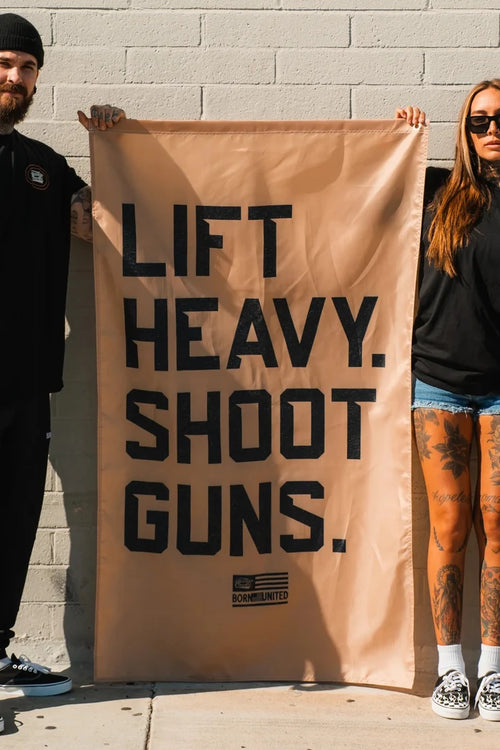 Two people stand against a light brick wall holding the LHSG - Flag by Tango Charlie Apparel, which reads “LIFT HEAVY. SHOOT GUNS.” Only their lower faces and bodies are visible.
