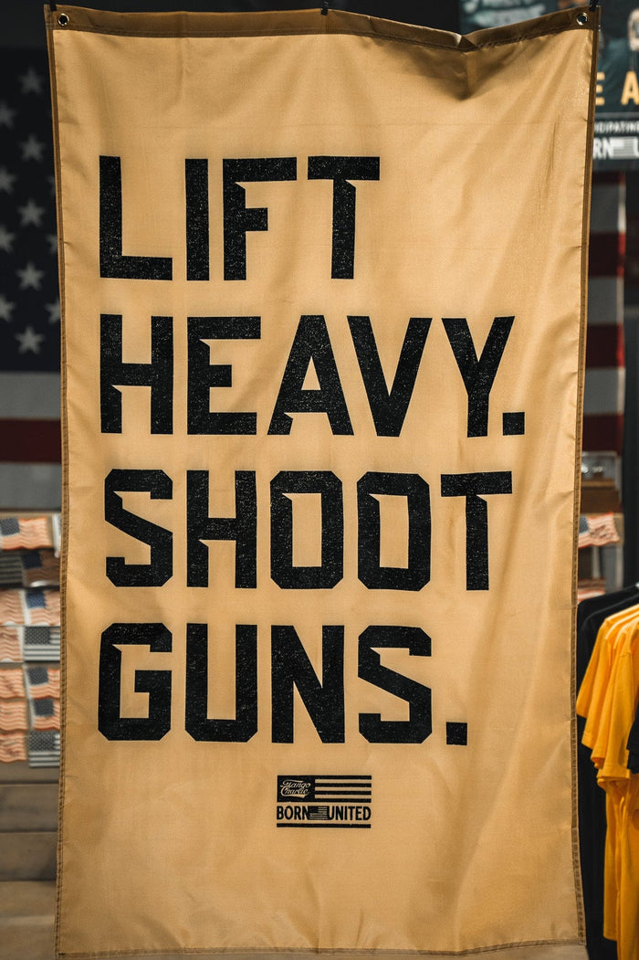A tan LHSG - Flag banner from Tango Charlie Apparel, with bold black text, hangs before a blurred backdrop of strength training shirts and a glimpse of a patriotic flag.