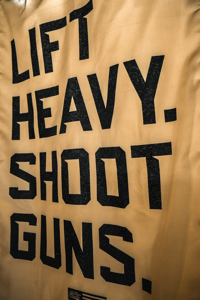 The LHSG - Flag by Tango Charlie Apparel is a tan banner featuring a patriotic flag and bold black vertical text, "LIFT HEAVY. SHOOT GUNS."—ideal for strength training or shooting enthusiasts.