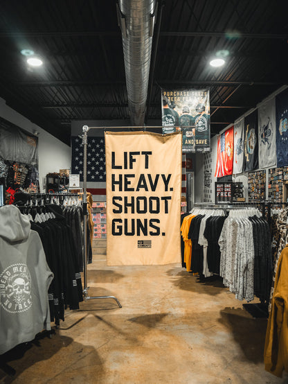 A banner reading "LIFT HEAVY. SHOOT GUNS." hangs above racks of Tango Charlie Apparel's strength training gear and the LHSG - Flag, with patriotic decor accenting the store’s warmly lit, industrial-style interior.