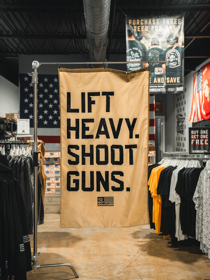 A large tan LHSG - Flag banner from Tango Charlie Apparel with bold black text hangs in a clothing store, surrounded by t-shirts and patriotic flag decor.