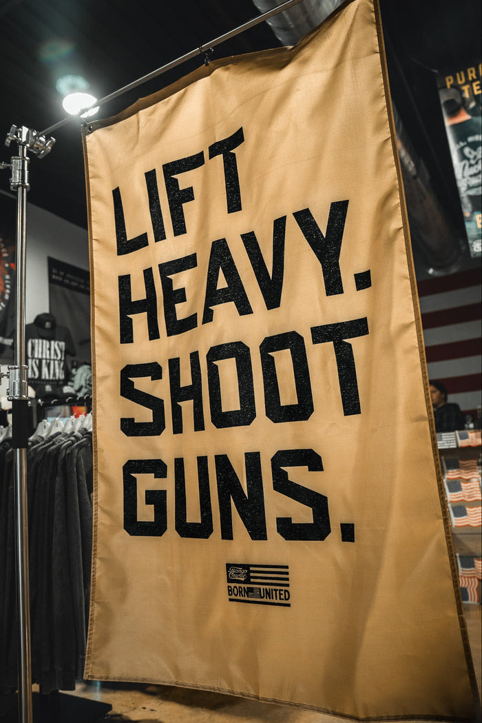 A large beige LHSG - Flag by Tango Charlie Apparel hangs in a store, featuring bold black LIFT HEAVY. SHOOT GUNS. text, merging strength and shooting themes. Clothing racks and posters are visible in the background.