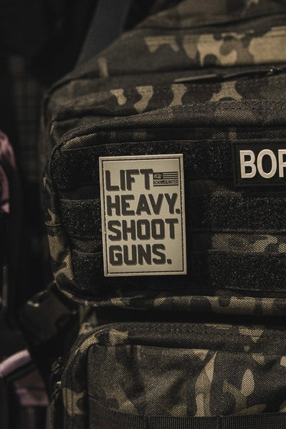 A close-up of a black camo backpack with the LHSG - Patch by Tango Charlie Apparel, featuring "LIFT HEAVY. SHOOT GUNS." in bold Velcro, alongside a 3D PVC patch reading "BORN" with an American flag. A pink bag appears on the left.