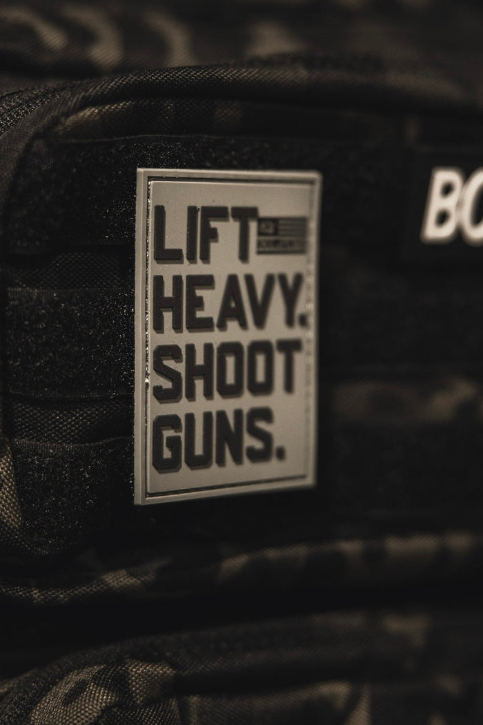 A close-up of a dark camouflage backpack shows a bold black 3D PVC Velcro LHSG - Patch by Tango Charlie Apparel that reads, LIFT HEAVY. SHOOT GUNS. Another blurred patch with partial text is seen above it.