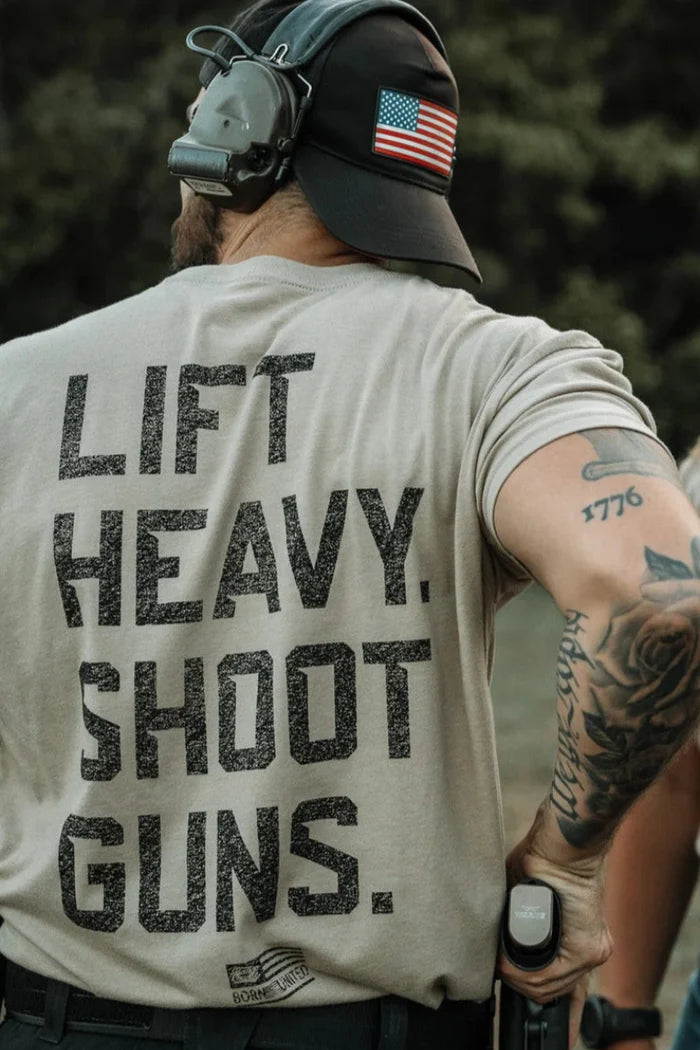 Wearing the Tango Charlie Apparel LHSG - Tan Tee, a person with tattoos on their right arm and a holstered handgun on their belt stands outdoors in high-intensity fitness gear and a backward cap with a US flag patch.