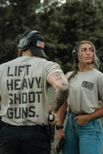 Two people outdoors wear LHSG - Tan Tee by Tango Charlie Apparel with “LIFT HEAVY. SHOOT GUNS.” on the man’s shirt. The woman holds a handgun, both sporting hearing protection amid trees, highlighting high-intensity fitness style.