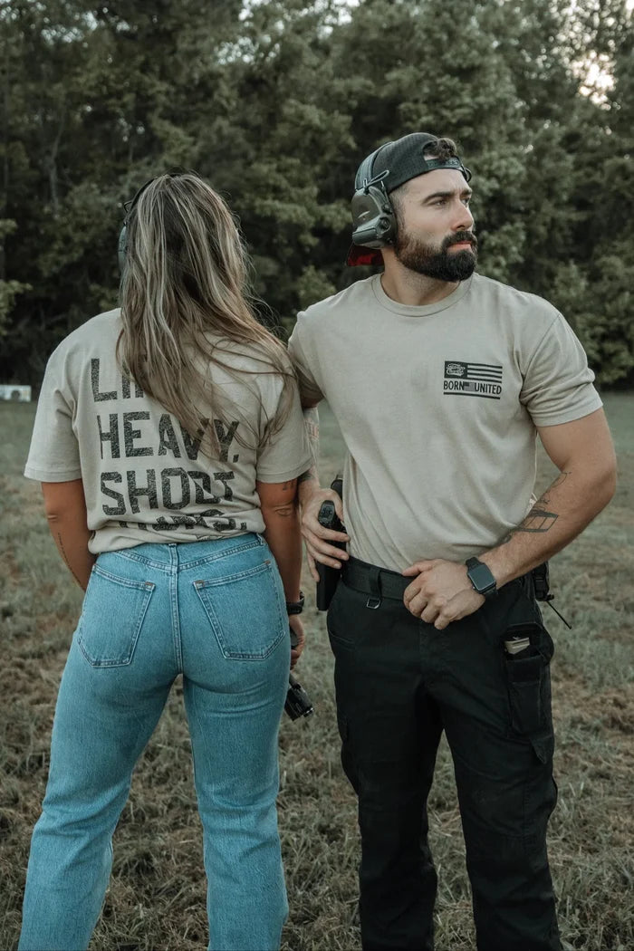 A woman and a man stand on grass, both wearing the LHSG - Tan Tee from Tango Charlie Apparel. They look relaxed with trees in the background.