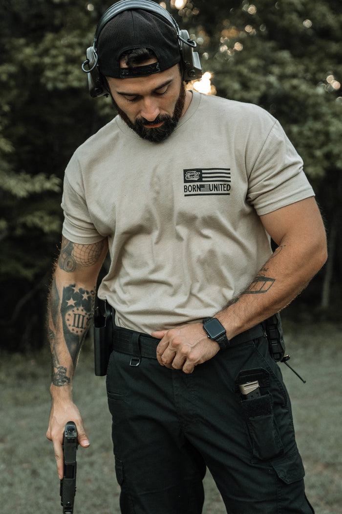 A bearded man in a backward cap and headphones, wearing a Tango Charlie Apparel LHSG Tan Tee and black cargo pants, stands outdoors holding a handgun with tattoos visible on his left arm.