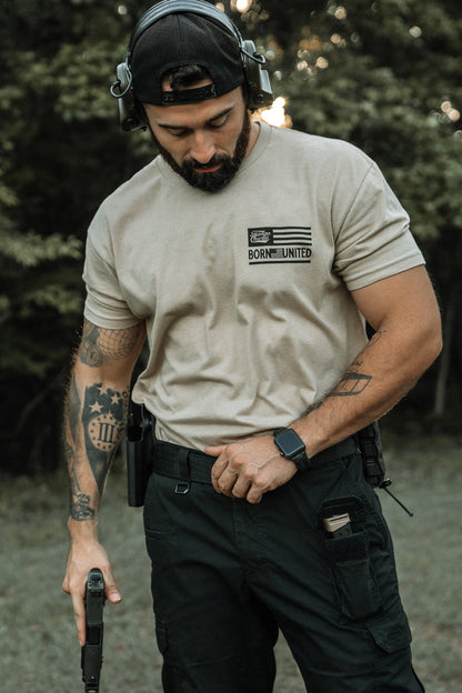 A bearded man in a backward cap and headphones, wearing a Tango Charlie Apparel LHSG Tan Tee and black cargo pants, stands outdoors holding a handgun with tattoos visible on his left arm.