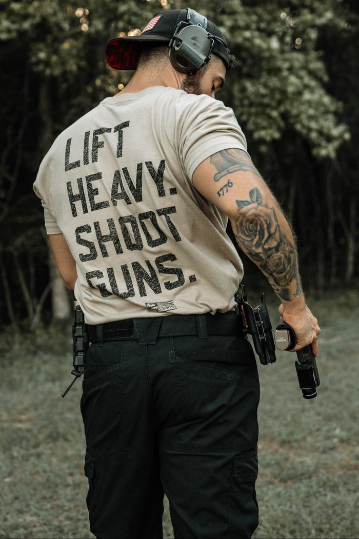 A man wearing the LHSG - Tan Tee by Tango Charlie Apparel stands outdoors in tactical pants, a cap, and hearing protection, gripping a handgun. His tattooed arm and gear-loaded belt complete his tactical look.