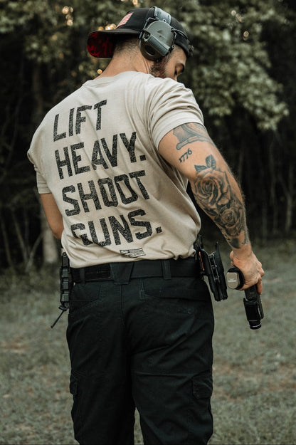 A man wearing the LHSG - Tan Tee by Tango Charlie Apparel stands outdoors in tactical pants, a cap, and hearing protection, gripping a handgun. His tattooed arm and gear-loaded belt complete his tactical look.