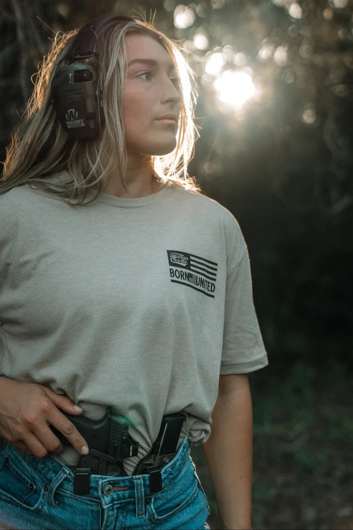 A woman wearing earmuffs and the LHSG - Tan Tee by Tango Charlie Apparel stands outdoors in sunlight, a holstered handgun tucked into her jeans, looking off to the side with a focused expression.