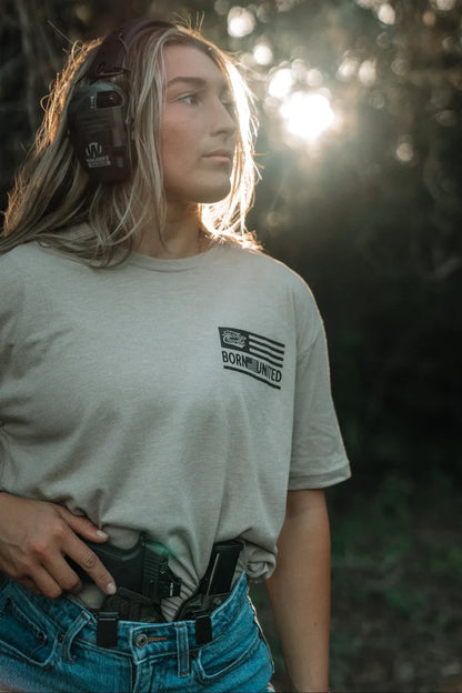 A woman wearing earmuffs and the LHSG - Tan Tee by Tango Charlie Apparel stands outdoors in sunlight, a holstered handgun tucked into her jeans, looking off to the side with a focused expression.