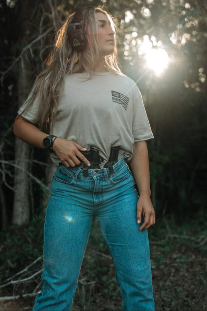 A woman with long hair stands outdoors in sunlight, wearing the LHSG - Tan Tee by Tango Charlie Apparel, headphones, and blue jeans. She rests her hand on a holstered gun, ready for action among the trees and greenery.