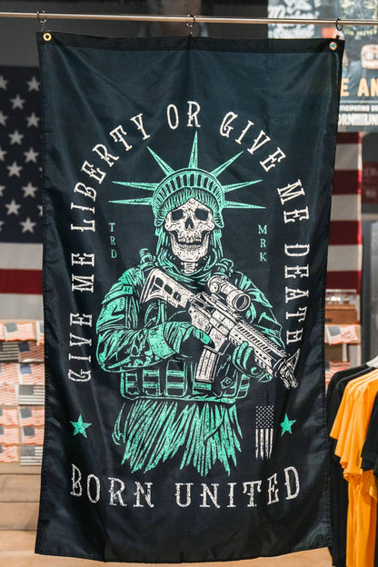 A Liberty or Death Flag by Born United Private Label, showing a skeleton in a Statue of Liberty crown with a rifle and the text “Give Me Liberty Or Give Me Death, Born United,” hangs in a clothing store with American flags and T-shirts in the background.