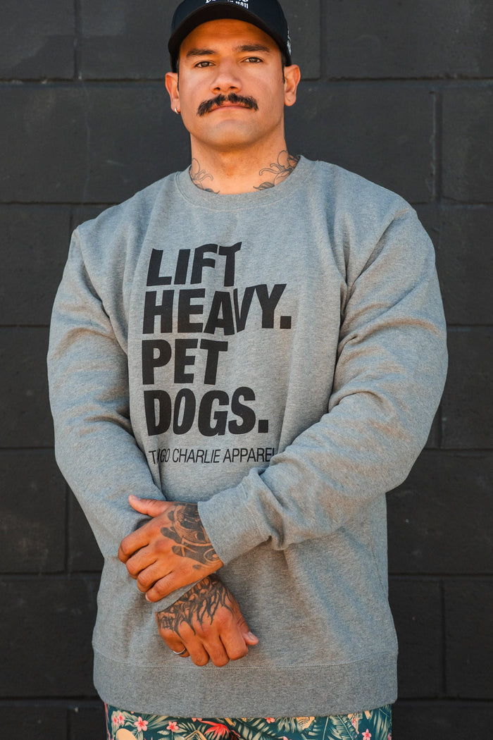A man wearing the Lift Heavy, Pet Dogs Crewneck by Tango Charlie Apparel stands against a black brick wall, arms crossed. He sports a mustache, tattoos, a black cap, and patterned shorts, showing his love for dogs.
