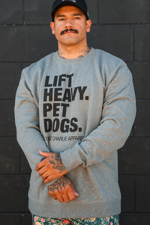 A man wearing the Lift Heavy, Pet Dogs Crewneck by Tango Charlie Apparel stands against a black brick wall, arms crossed. He sports a mustache, tattoos, a black cap, and patterned shorts, showing his love for dogs.