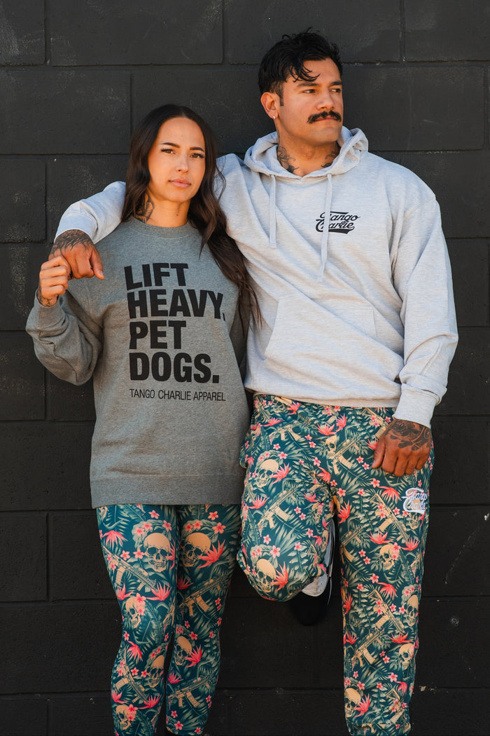 A woman wears Tango Charlie Apparel's "Lift Heavy. Pet Dogs Crewneck," standing in colorful floral leggings beside a man in a light gray hoodie, both against a black brick wall. His arm rests on her shoulder.