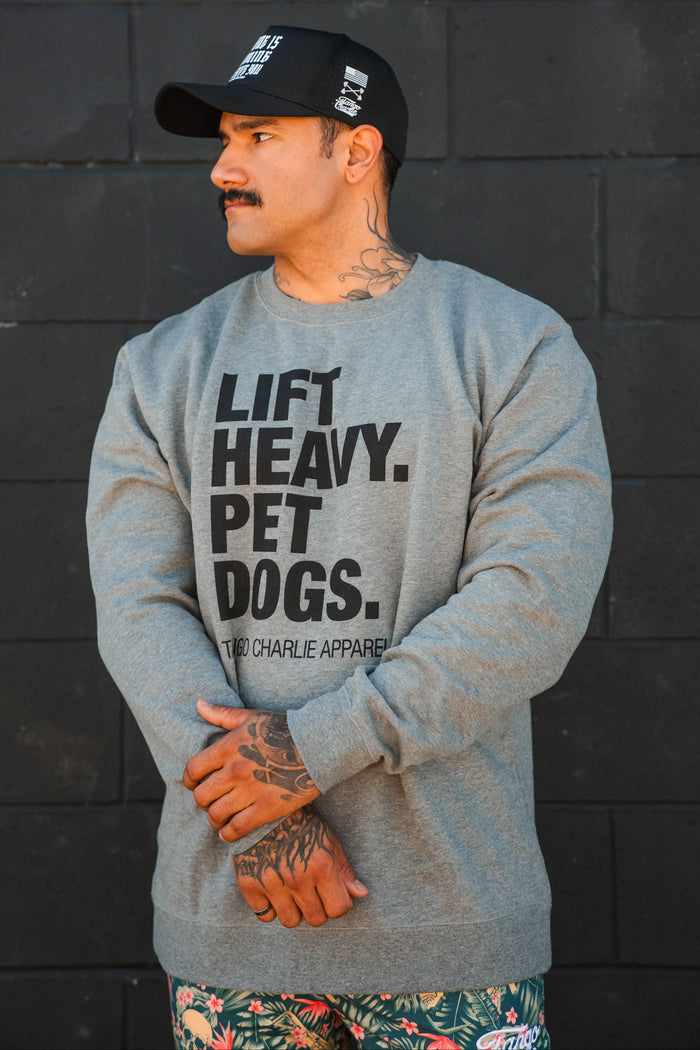 A tattooed man stands arms crossed before a black brick wall, wearing Tango Charlie Apparel’s Lift Heavy. Pet Dogs Crewneck and floral shorts, his black cap and dog-loving vibe making the statement on his gray sweatshirt clear.