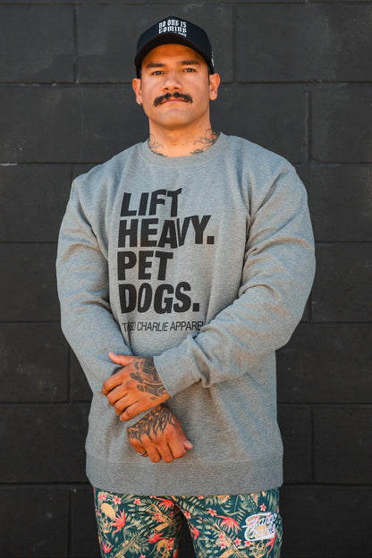 A man with a mustache stands against a black wall, wearing a black cap, colorful floral shorts, and the Lift Heavy. Pet Dogs Crewneck by Tango Charlie Apparel. He has hand tattoos and looks like someone who truly loves dogs.