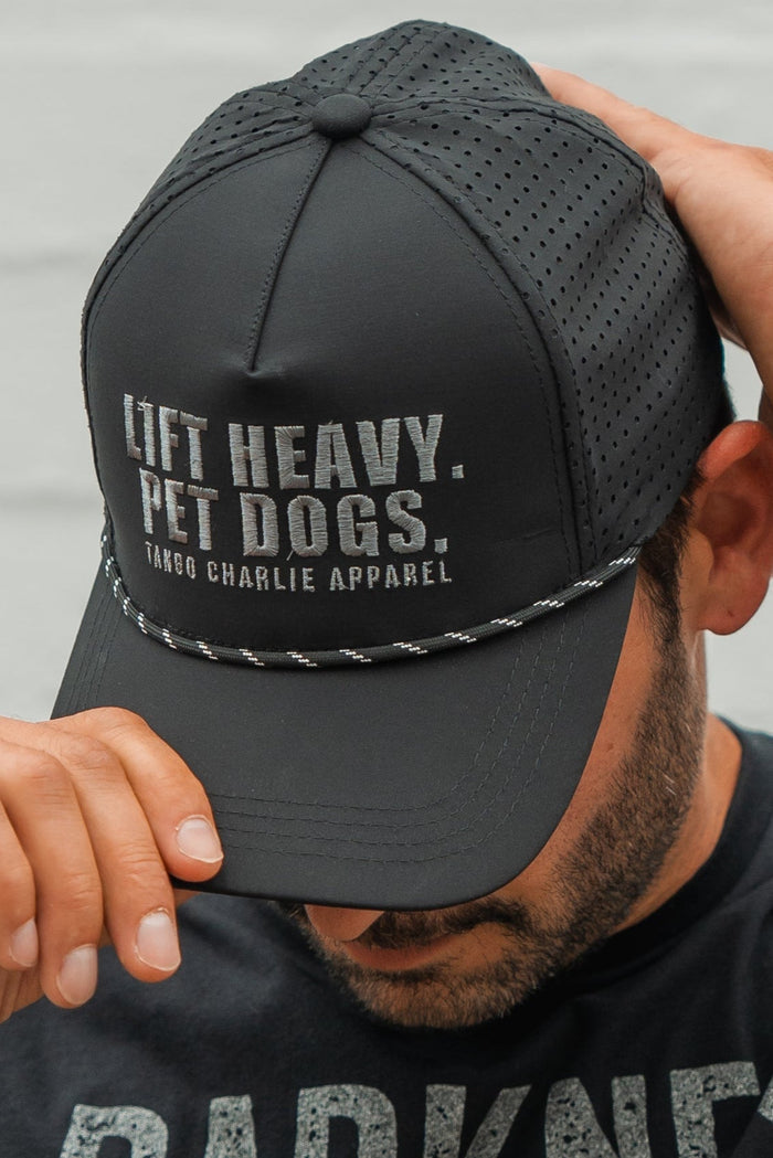 A man in a black shirt, sporting a trimmed beard, adjusts the Tango Charlie Apparel Lift Heavy. Pet Dogs Snapback—a black, water-resistant cap with perforated panels, braided cord, and bold white text: LIFT HEAVY. PET DOGS.