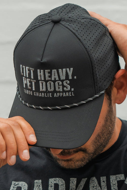A man in a black shirt, sporting a trimmed beard, adjusts the Tango Charlie Apparel Lift Heavy. Pet Dogs Snapback—a black, water-resistant cap with perforated panels, braided cord, and bold white text: LIFT HEAVY. PET DOGS.