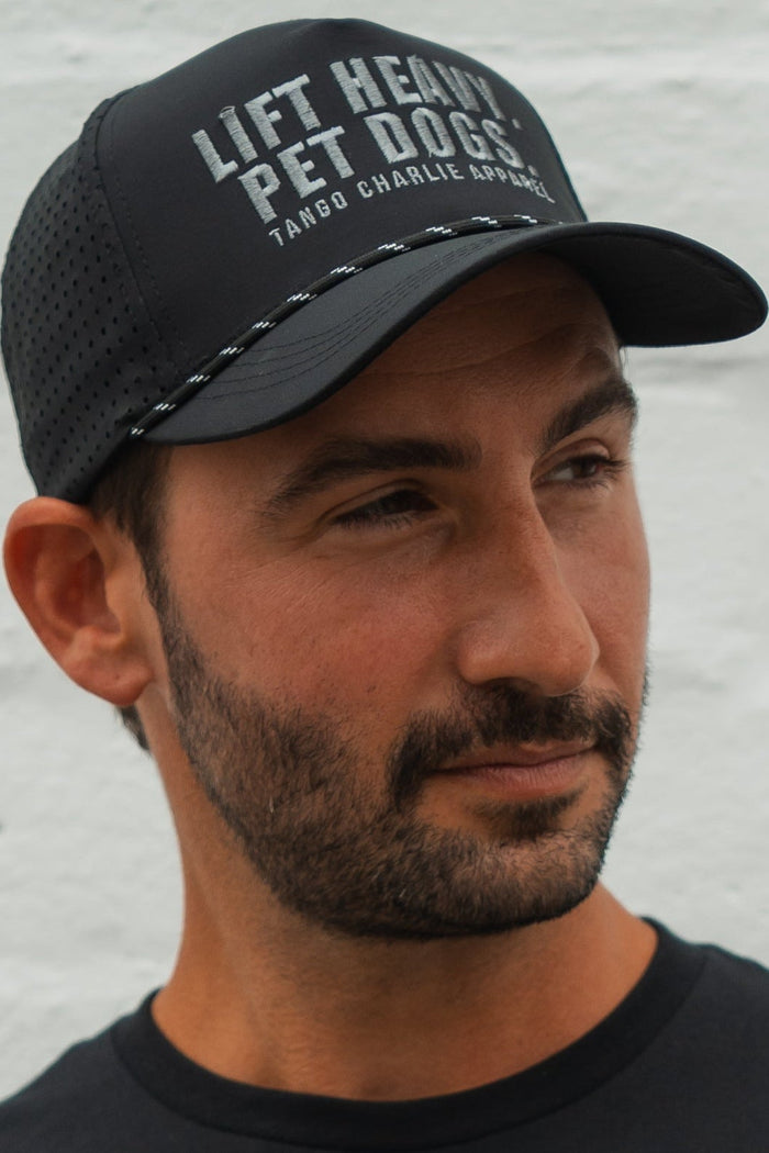 A bearded man wears the Tango Charlie Apparel Lift Heavy. Pet Dogs Snapback, a black, water-resistant hat with the slogan on front, as he looks to the side against a light background—ideal for lifting and dog lovers.