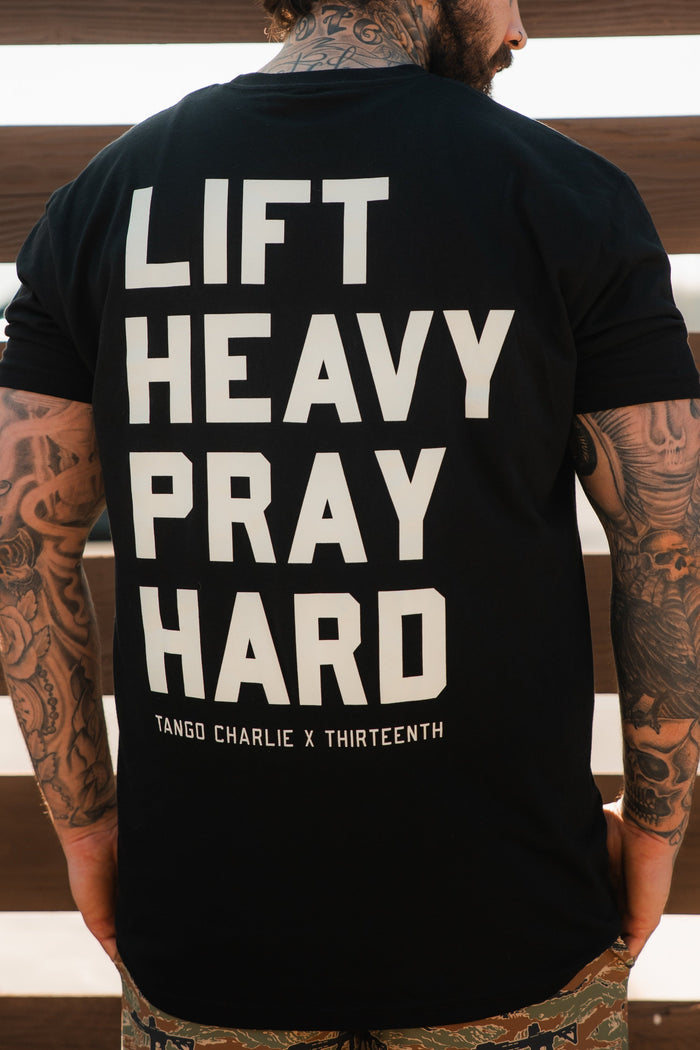 A tattooed man is seen from behind wearing the Lift Heavy. Pray Hard. - Black Tee by Tango Charlie Apparel, featuring bold white text and merging faith with fitness style.