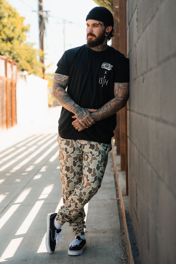 A tattooed man in a black beanie, Tango Charlie Apparel "Lift Heavy. Pray Hard." black tee, camouflage pants, and black sneakers leans casually against a concrete wall on a sunny sidewalk.