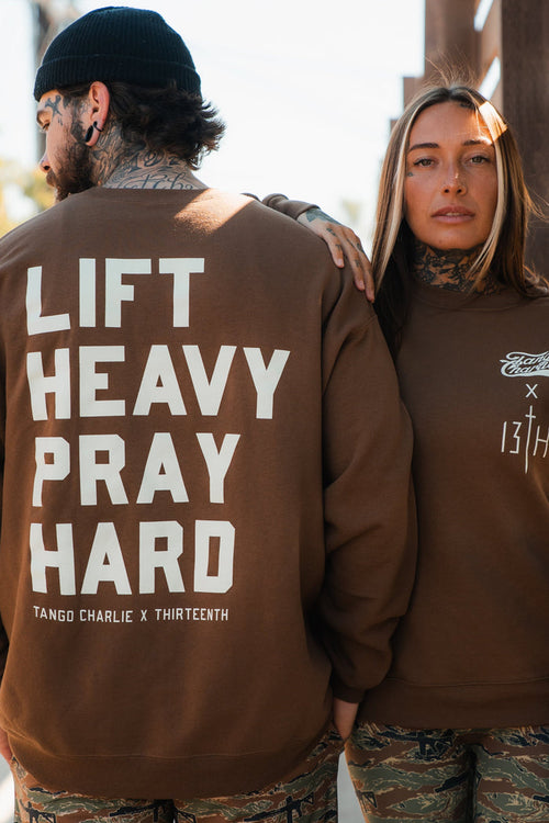 Two tattooed people in brown Tango Charlie Apparel crewnecks and camo pants stand outdoors. One sweatshirt reads "Lift Heavy. Pray Hard." on the back, while the other displays Tango Charlie x Thirteenth logos on the chest.