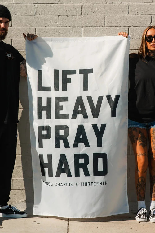 Two people stand against a brick wall, holding up the Lift Heavy. Pray Hard. - Flag from Tango Charlie Apparel. The bold banner reads, “LIFT HEAVY PRAY HARD,” making a powerful statement about faith and fitness. Both individuals have tattoos and are dressed in dark clothing.