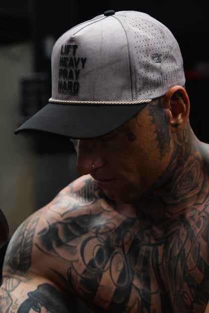 A tattooed individual wearing the Lift Heavy. Pray Hard. Deadwood Camo Hydro Snapback Hat by Tango Charlie Apparel in grey, gazing downward in a dimly lit environment.