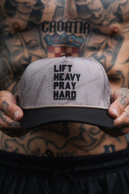 A shirtless, tattooed person holds a gray and black Tango Charlie Apparel “Lift Heavy. Pray Hard. Deadwood Camo Hydro” snapback hat with the words “LIFT HEAVY PRAY HARD” printed on the front.