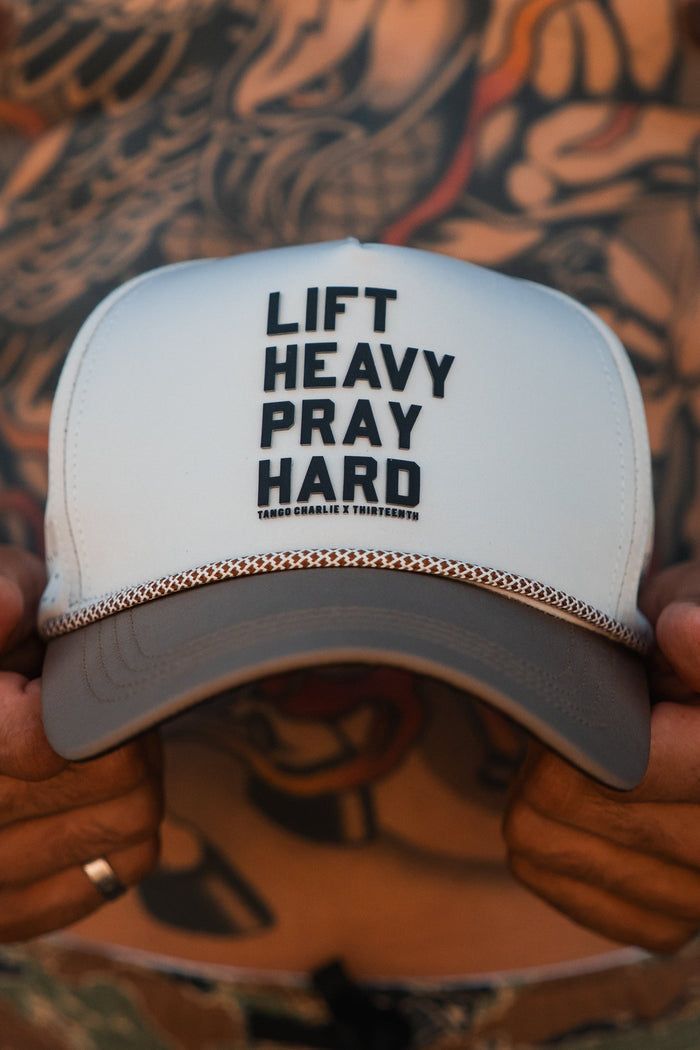 A person with heavily tattooed arms and torso holds the "Lift Heavy. Pray Hard. Hydro - Snapback Hat" by Tango Charlie Apparel, a white and brown gym hat featuring bold front text.