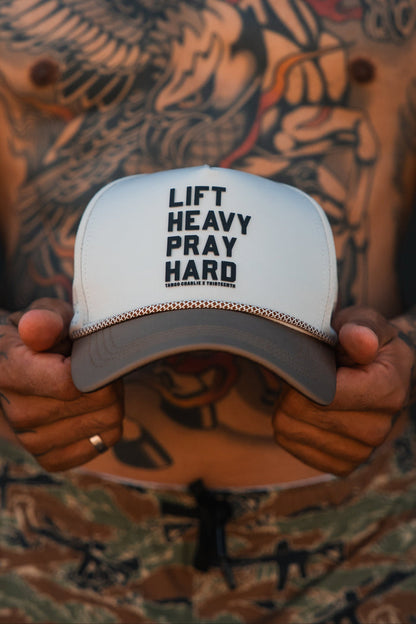 A tattooed individual wearing camouflage pants and a ring holds the Tango Charlie Apparel Lift Heavy. Pray Hard. Hydro Snapback Hat, showcasing this stylish and practical gym hat perfect for tough workouts.