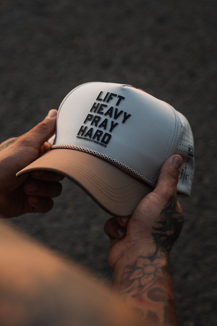 A person with tattooed forearms holds the white and beige "Lift Heavy. Pray Hard. Hydro - Snapback Hat" by Tango Charlie Apparel, featuring bold front text. The blurred asphalt background highlights this stylish gym hat.
