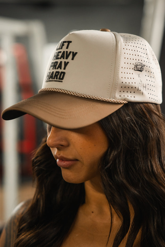 A woman with long dark hair wears the Lift Heavy. Pray Hard. Hydro Snapback Hat by Tango Charlie Apparel, featuring bold black lettering. The background is blurred, suggesting she's in an indoor gym setting.