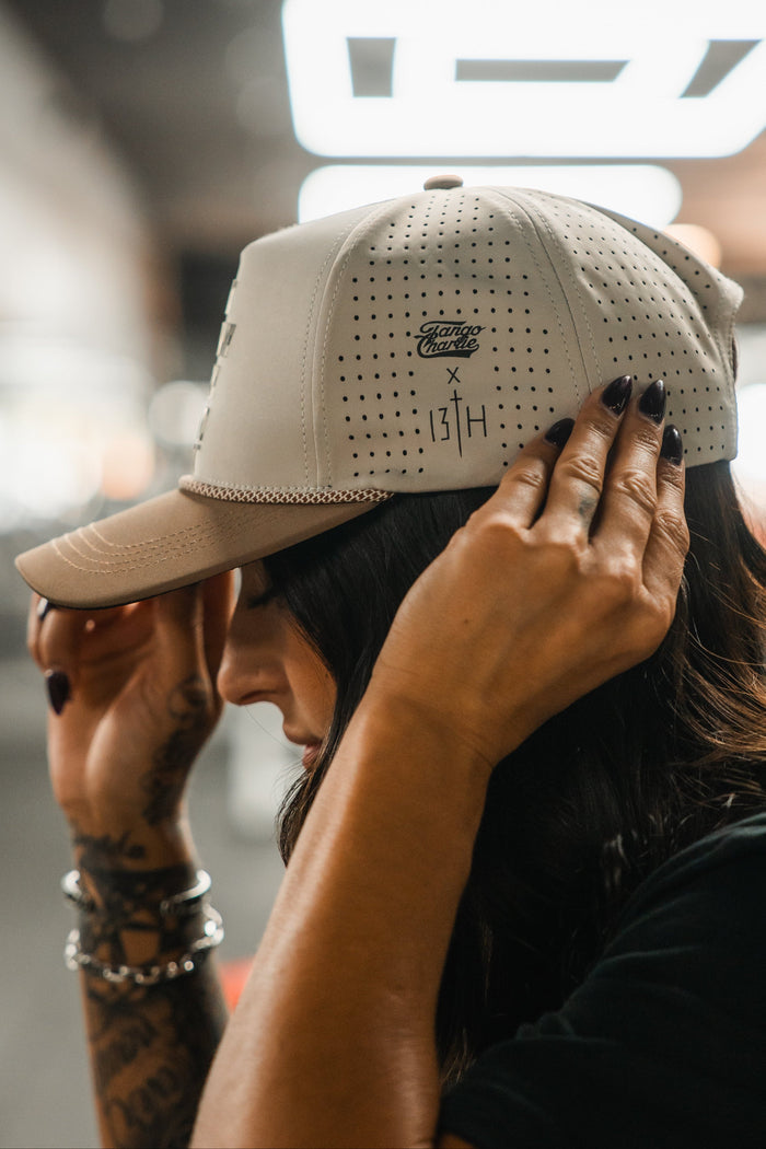 A person with long dark hair and tattoos adjusts a light-colored, perforated Lift Heavy. Pray Hard. Hydro - Snapback Hat by Tango Charlie Apparel, showcasing bracelets and dark-painted nails.