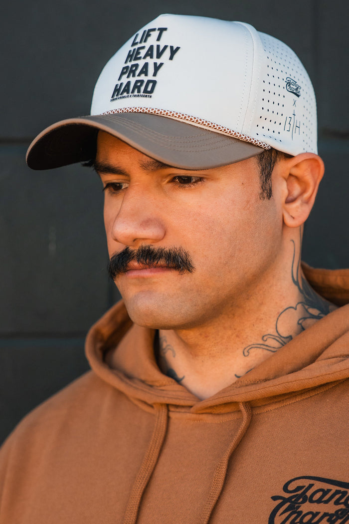 A man with a mustache, neck tattoos, and a tan hoodie looks down thoughtfully. He wears the Tango Charlie Apparel Lift Heavy. Pray Hard. Hydro Snapback Hat against a dark background.
